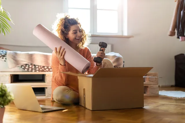 A woman at home unpacking a delivered box containing a yoga mat and hand weight